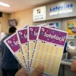 Mão segurando vários volantes da Lotofácil dentro de uma unidade das Loterias Caixa.