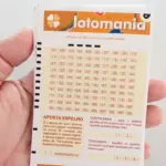 Mão segurando um volante de apostas da Lotomania em papel branco com o logotipo oficial laranja, mostrando a grade de números de 01 a 00 e os campos de Aposta Espelho, Surpresinha e Teimosinha.