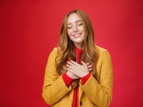 girl feeling like heart warming as remembering sweet tender memories with closed eyes silly broad smile holding hands chest pressing soul posing nostalgic touched red background min 1