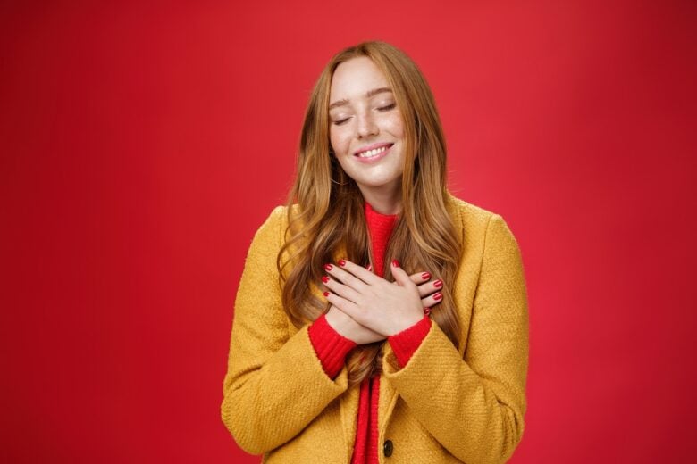 girl feeling like heart warming as remembering sweet tender memories with closed eyes silly broad smile holding hands chest pressing soul posing nostalgic touched red background min 1