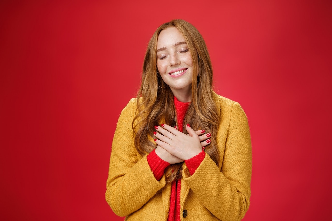 girl feeling like heart warming as remembering sweet tender memories with closed eyes silly broad smile holding hands chest pressing soul posing nostalgic touched red background min 1