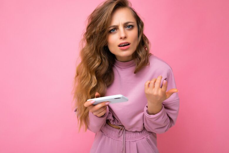 Bela jovem loira pensativa vestindo moletom rosa isolado sobre fundo rosa com cópia espaço segurando e usando telefone celular olhando para a câmera e tendo perguntas - Fotos do Canva