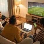 Pessoas assistindo ao jogo Al-Taawoun x Al-Ittihad em uma TV, analisando estatísticas em um laptop e celular.