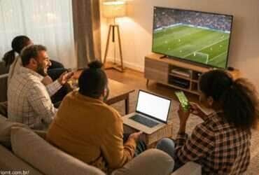 Pessoas assistindo ao jogo Al-Taawoun x Al-Ittihad em uma TV, analisando estatísticas em um laptop e celular.