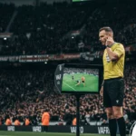 Foto de árbitro consultando o VAR durante partida de futebol em estádio lotado.