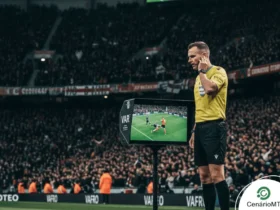 Foto de árbitro consultando o VAR durante partida de futebol em estádio lotado.