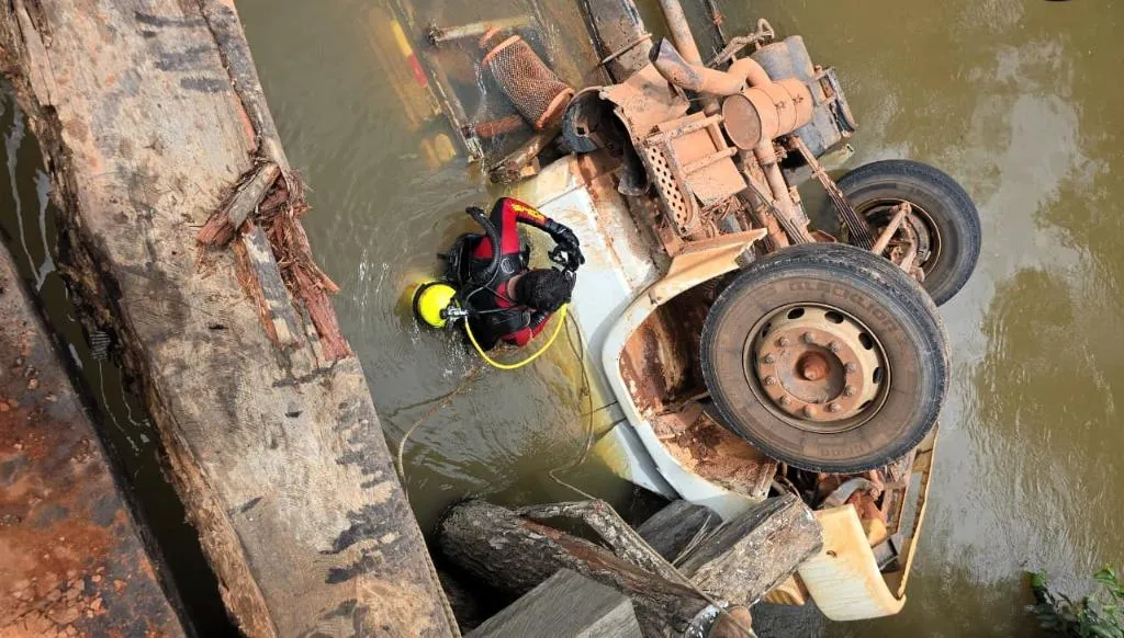 Caminhão de carga tombado de rodas para cima e parcialmente submerso em um rio, com um mergulhador do Corpo de Bombeiros com roupa vermelha e preta realizando o resgate.