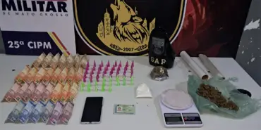 Materiais apreendidos pela Polícia Militar em Várzea Grande, incluindo diversas cédulas de Real, pinos coloridos de cocaína, porções de maconha, balança de precisão, rolos de plástico filme e um smartphone sobre uma mesa branca.