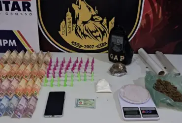 Materiais apreendidos pela Polícia Militar em Várzea Grande, incluindo diversas cédulas de Real, pinos coloridos de cocaína, porções de maconha, balança de precisão, rolos de plástico filme e um smartphone sobre uma mesa branca.
