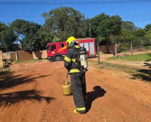 A imagem mostra um bombeiro bem equipado em frente a um botijão de gás. Um caminhão do corpo de bombeiros pode ser visto ao fundo