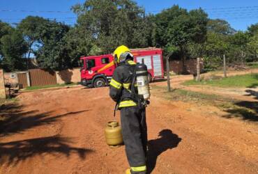 A imagem mostra um bombeiro bem equipado em frente a um botijão de gás. Um caminhão do corpo de bombeiros pode ser visto ao fundo