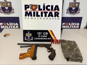 Foto das armas apreendidas em Canarana, as armas estão organizadas em uma mesa em frente à um banner com a logo da Polícia Militar de Mato Grosso