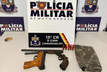 Foto das armas apreendidas em Canarana, as armas estão organizadas em uma mesa em frente à um banner com a logo da Polícia Militar de Mato Grosso