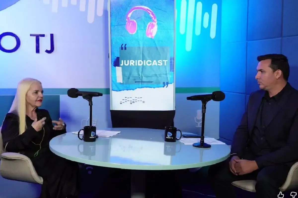 Uma mulher de cabelos longos e claros, vestindo preto, e um homem de terno estão sentados em lados opostos de uma mesa redonda azul em um estúdio de podcast, com microfones e uma tela vertical ao fundo