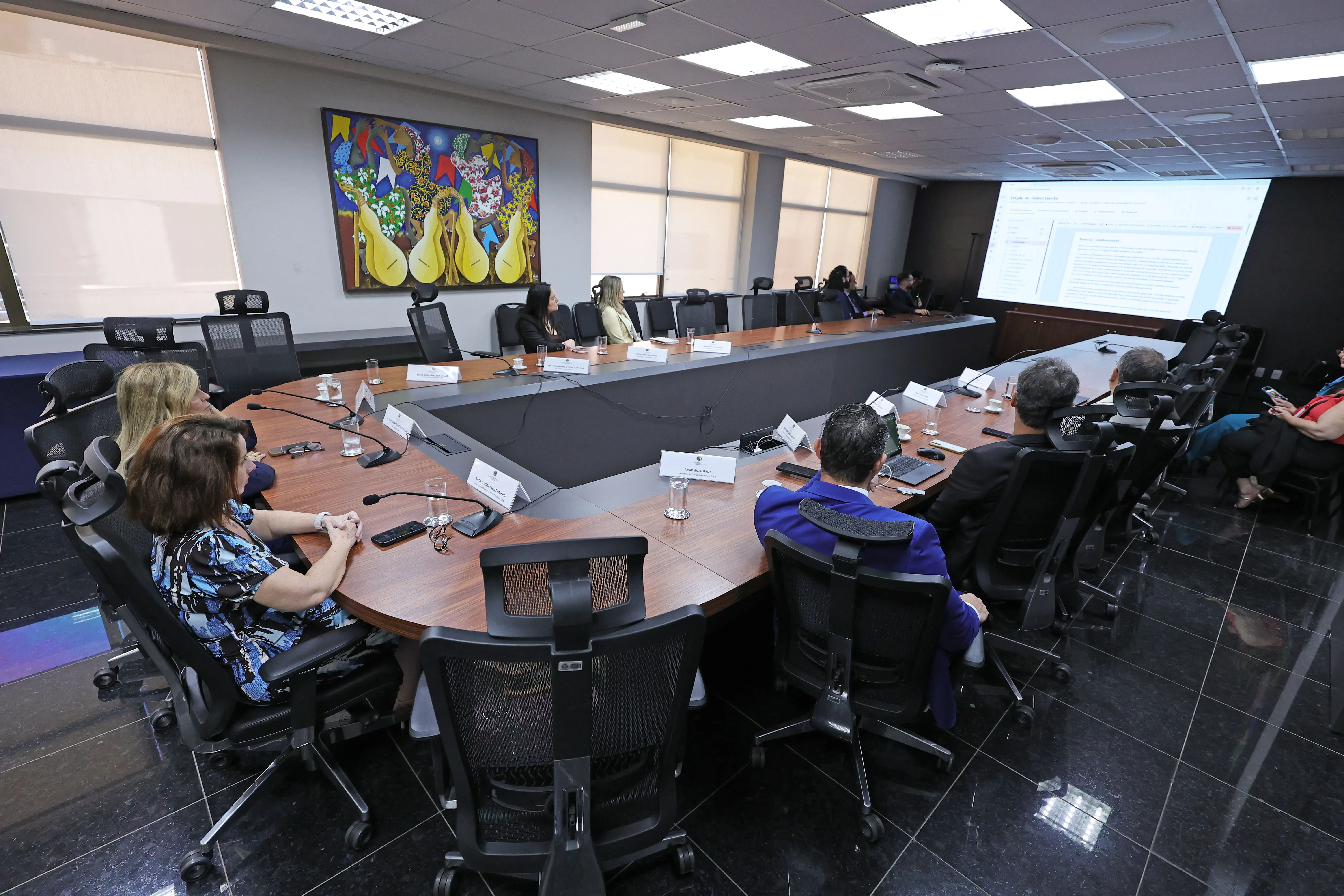 Ampla sala de reuniões com mesa oval, diversas pessoas sentadas em cadeiras pretas acompanham apresentação em telão. Ao fundo, quadro colorido decora a parede e janelas com persianas claras.