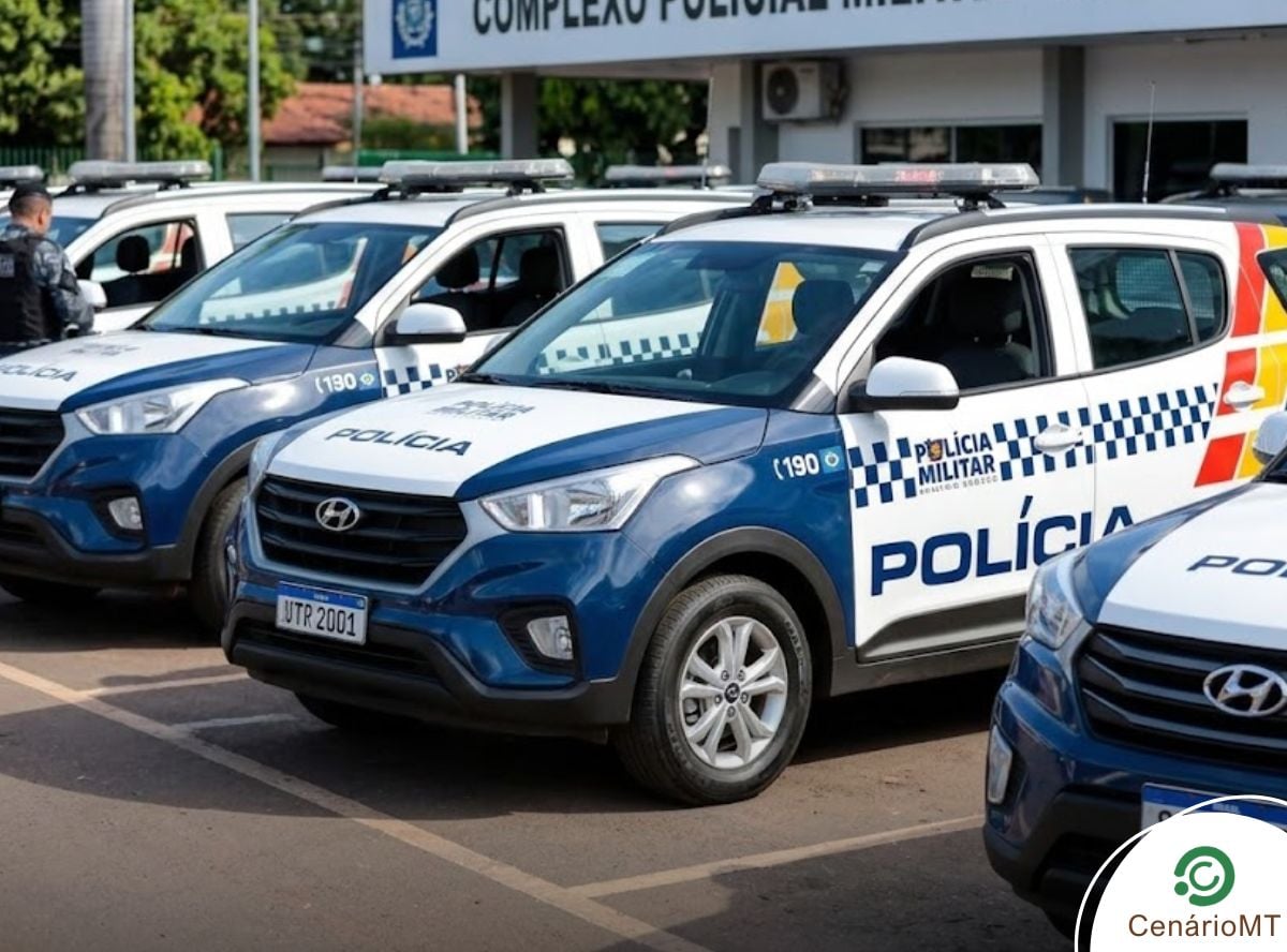 Uma frota de picapes brancas e azuis da Polícia Militar de Mato Grosso (PMMT) estacionada em uma linha ordenada em um pátio pavimentado durante o dia. Os veículos apresentam a identidade visual oficial, incluindo as listras chevron vermelhas e laranjas nas portas e as palavras 'POLÍCIA MILITAR' e 'MATO GROSSO' em destaque. O edifício de comando ao fundo tem uma placa grande que diz 'POLÍCIA MILITAR - MATO GROSSO'. Dois oficiais uniformizados são visíveis perto dos veículos.