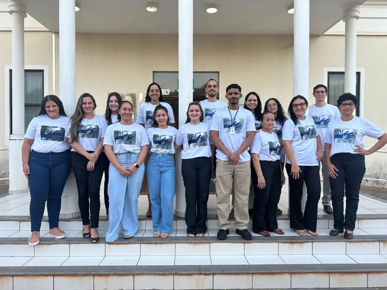 Grupo de pessoas sorri alinhado nas escadas da entrada de um prédio. Todas vestem camisetas brancas padronizadas com a estampa da campanha "Pai Presente".