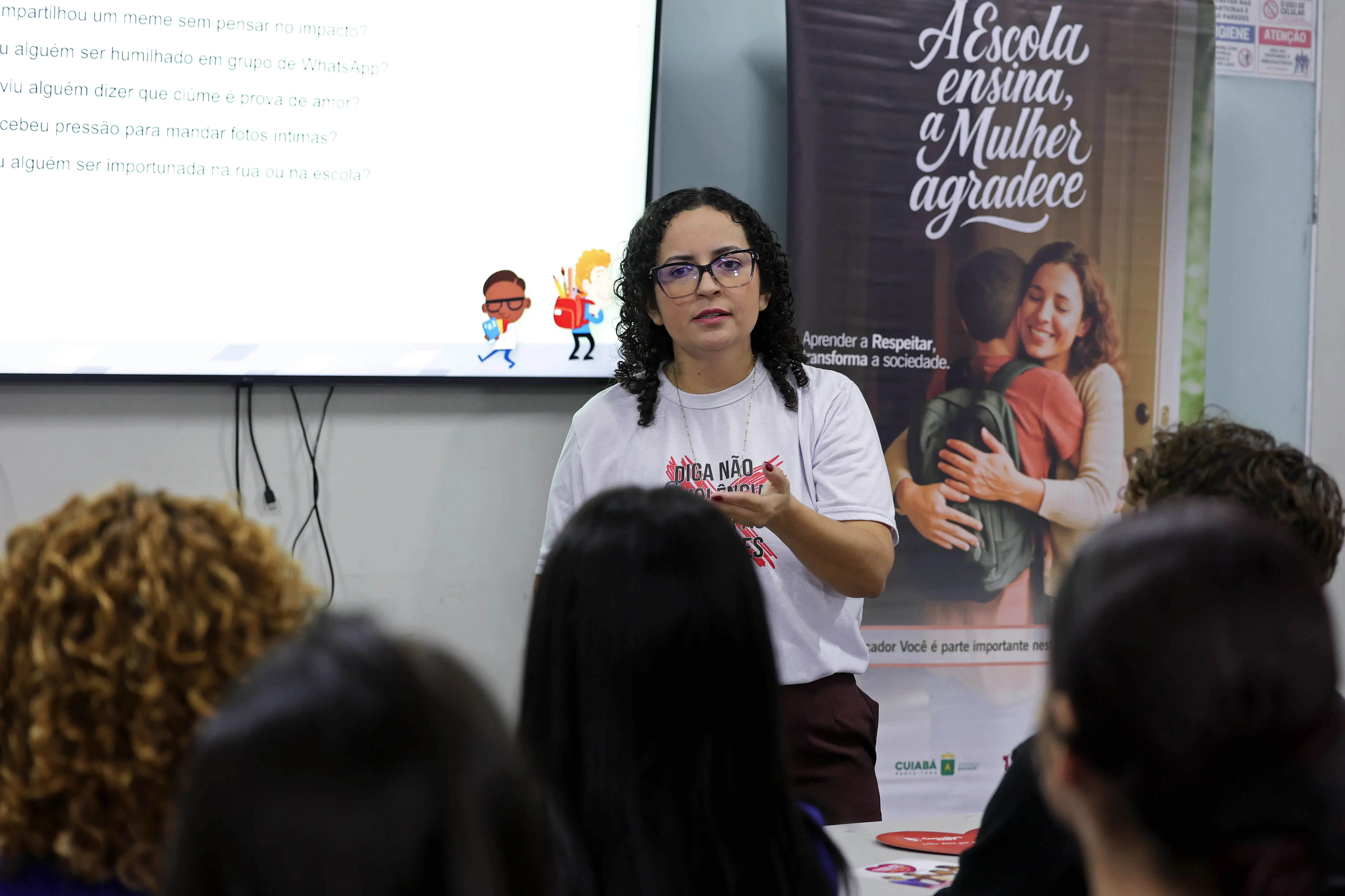 Mulher de óculos, cabelos cacheados e camiseta branca gesticula ao falar. Ao fundo, vê-se uma tela com perguntas e um banner com o texto "A Escola ensina, a Mulher agradece".