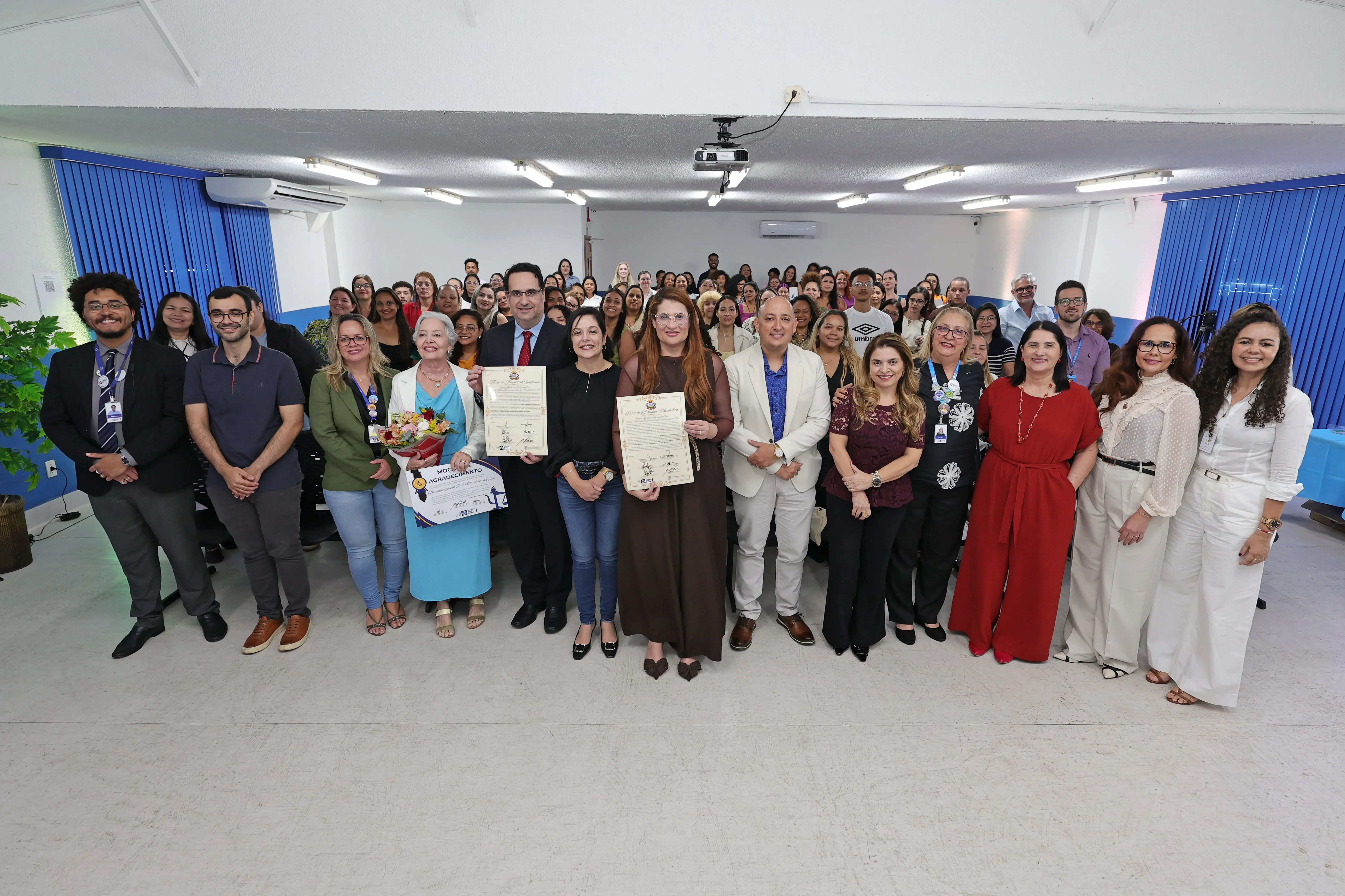 Grupo de facilitadores e autoridades posa para foto em auditório. À frente, um homem de terno escuro e uma mulher de vestido marrom exibem certificados emoldurados recebidos durante o evento.