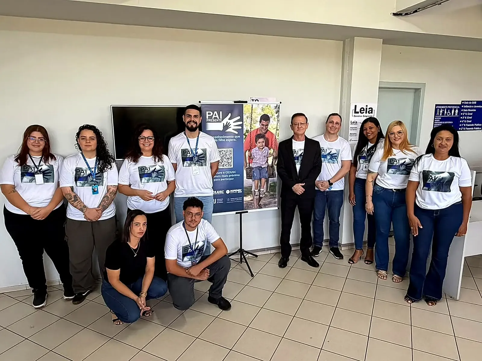 Servidores usam camisetas brancas do projeto "Pai Presente" e posam para foto em sala do fórum, junto a banner azul do CEJUSC. Ao centro, homem de terno escuro acompanha o grupo